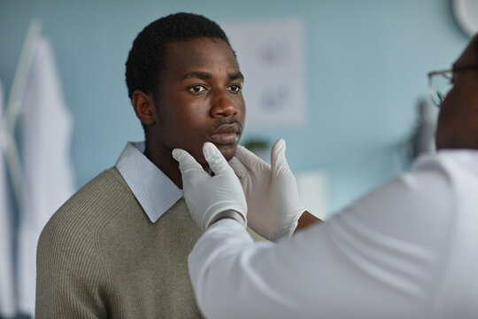 Black teenage boy receiving medical examination from male doctor wearing gloves, doctor gently touching boys chin while boy looking forward, clinical setting visible in background