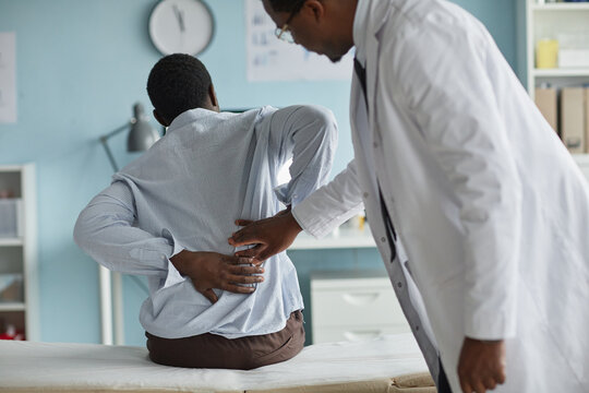 Black young adult man sitting on examination table holding lower back while Black male doctor in coat examining spine during medical consultation in clinic