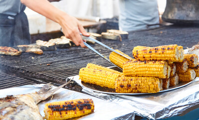 Hands of chef use tongs to place grilled corn on cob on plate, with pieces of fried fish in background. Concept: street food, healthy eating, urban space