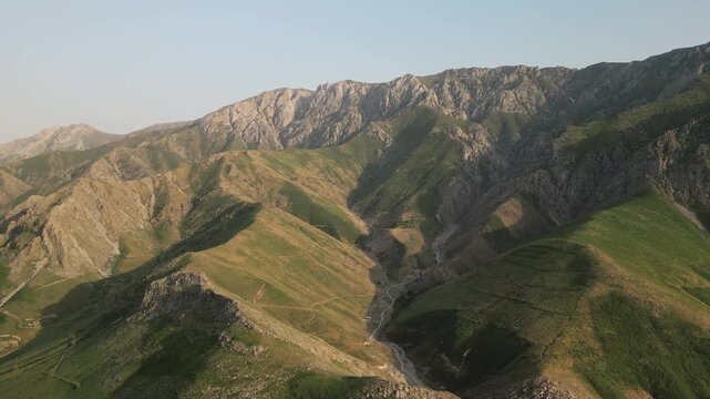 Aerial view across a wide mountain basin in the Baysuntau range, Boysun district. Dynamic light sweeps over ridge and gorge, highlighting travel route, dirt road and vast summer scenery