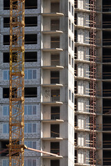 Vertical close-up view of modern apartment tower under construction with yellow crane, exposed concrete, brick walls, scaffolding, and blue windows.