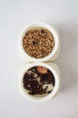 Two white square bowls containing spices, including white peppercorns and cloves, displayed against a white background