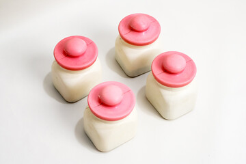 Four small square jars with white content and a pink hat-shaped lid, placed on white