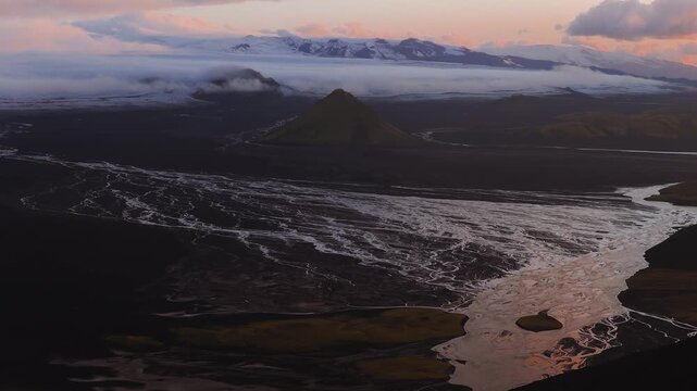 Sweeping aerial view of Maelifell volcano, rivers, volcanic terrain, glacier, and mountains under warm sunset hues with low lying clouds in Iceland.