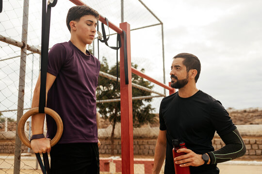 Teenager practicing calisthenics on gymnastic rings with adult supervision at outdoor park
