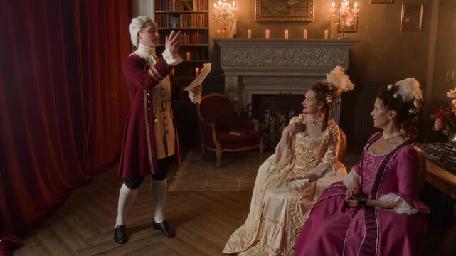 Full shot of young 19th century Caucasian gentleman, dressed in red frock coat, breeches, stockings, wig reciting own love poems to two noble ladies in fashionable historical costumes
