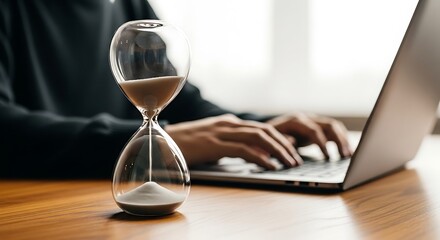 Hourglass measuring time on a desk with a person working on a laptop