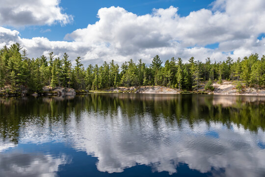 Beautiful Reflection of Blue Sky, White Clouds, Evergreen and Pink Granite Rocks on Moira Pond, French River, Ontario