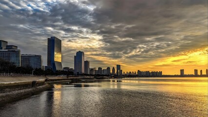 Obraz premium Modern City Skyline Reflected in Calm Waters During Dramatic Golden Sunrise with Cloudy Sky