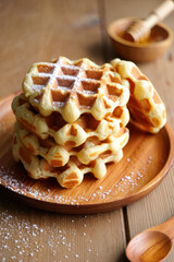 Golden Belgian waffles stacked on a wooden plate, sprinkled with powdered sugar, with a honey dipper and spoon nearby, cozy breakfast scene on a rustic wooden table