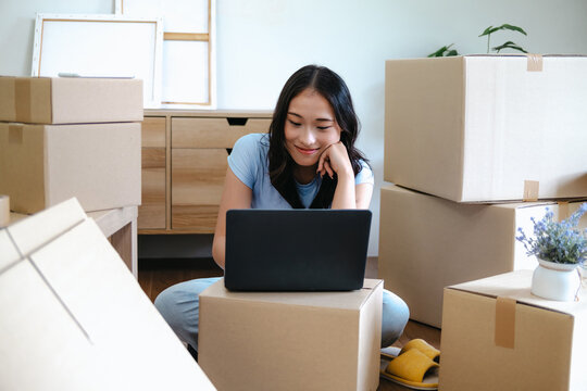 Asian woman using laptop among moving boxes preparing for home relocation