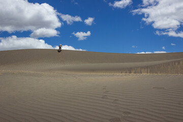 A single person stands on the crest of a large sand dune in Palmira, Ecuador, with a vibrant blue sky and white clouds above. Adventure, exploration, and solitude in a vast landscape.