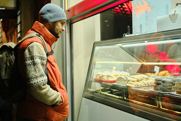 A man in a red vest and backpack looks at pastry display in a bakery window at night
