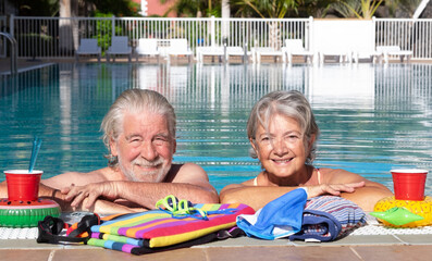 Happy senior couple relaxing together at a swimming pool, smiling and embracing at the poolside. Colorful towels and floating drink holders create a cheerful summer vacation atmosphere