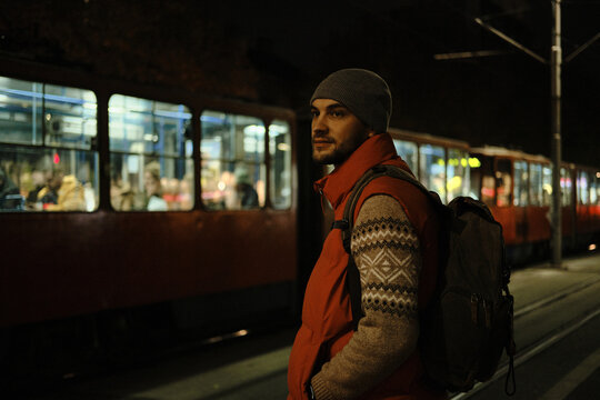 A man in a red vest stands near a tram stop at night with trams and people in the background