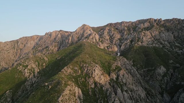 Aerial view of sunlit Chimgan ridge in the Ugam-Chatkal mountains, Uzbekistan. Golden hour reveals rocky crest, alpine meadow and seasonal snow line, inviting hiking, tourism and drone cinematography