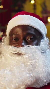 Santa Claus portrait thums up putting gifts under christmas tree in living room, making surprise. African american man in costume of Santa puts present box for children. Saint Nicholas working at