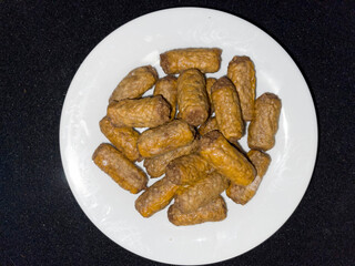 This is a high-contrast, top-down photograph of a pile of small, golden-brown, finger-shaped fried food items or sausages, tightly clustered on a plain white plate against a black background.