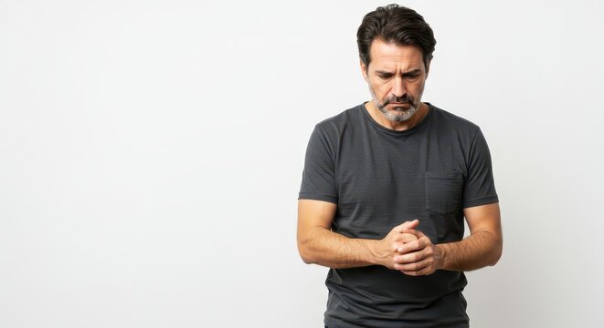 A middle-aged man, with a thoughtful expression, stands against a plain background, hands clasped together, conveying a feeling of contemplation or sadness.