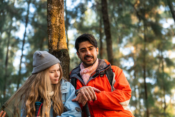 Fototapeta premium Young Couple Resting Against Tree During a Winter Hike. Enjoying the Golden Hour Light in a Peaceful Forest, Wearing Warm Clothes and Backpacks, Sharing a Quiet and Happy Moment Together
