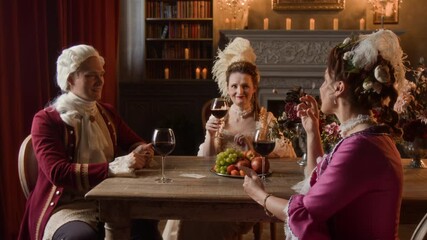 Medium full shot of young 19th century Caucasian gentleman in red frock coat, wig and two mature ladies in fashionable dresses sitting at table in manor parlor, drinking wine with fruits and chatting - Powered by Adobe