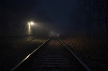 A foggy railway at night is dimly lit by wall mounted lights, casting shadows along empty tracks. The quiet and mysterious mood evokes suspense and solitude.