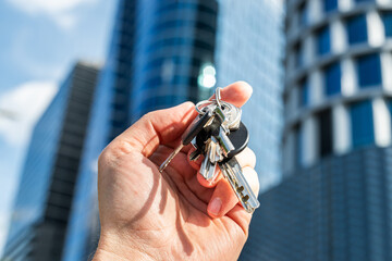 Real Estate Keys Held by Hand in Front of Modern Office Building. Close-up shot of a hand holding several metal keys in front of a sleek modern glass skyscraper