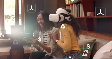 Two women using tablet and VR headset on sofa, adjusting energy simulation icons for renewables - Powered by Adobe