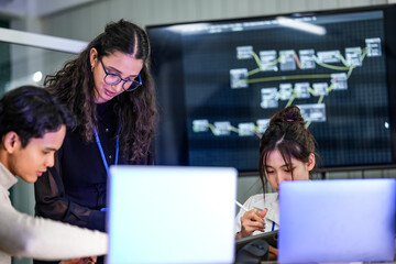 A female team leader mentors her junior IT staff in a tech office. The diverse group is working together on a complex software project, analyzing data on their computers.