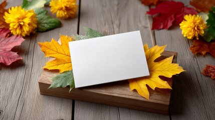 A white card is on a wooden surface with autumn leaves and flowers. The card is blank, but it could be used for a variety of purposes, such as a thank you note, a reminder