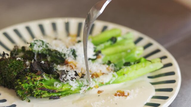 Close-up of roasted broccolini with parmesan and crunchy crumbs on ceramic plate. Selective focus. Fork pressing vegetables. Healthy, vegetarian, flavorful dish captured in natural warm lighting.