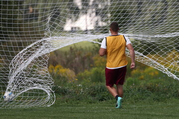 Man in orange t-shirt and brown shorts in front of football goal with scored ball