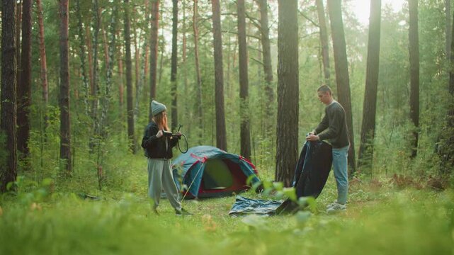 Young woman holding hose stands in forest clearing watching man spray tent fabric near set-up tent, both surrounded by tall trees and morning sunlight in calm outdoor camping environment