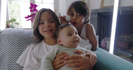 Siblings with baby on couch, girl holding baby, boy making funny face with ping pong ball, family...