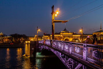Fototapeta premium Evening view of the Palace Bridge over the Neva River with glowing lanterns, ornate iron railing, and reflections of city lights. Saint Petersburg, Russia.