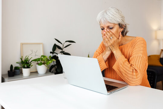 Shocked senior adult woman receiving bad news on laptop computer message sitting on table at home - Powered by Adobe