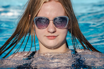 Teenage girl relaxing in a pool with stylish sunglasses and wet hair, enjoying a sunny summer day surrounded by cool blue water