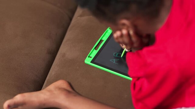 African girl child writing the alphabets ABCD on the screen of a digital drawing tablet at home.