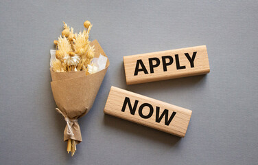 Apply now symbol. Concept word Apply now on wooden blocks. Beautiful grey background with bouquet of flowers. Business and Apply now concept. Copy space