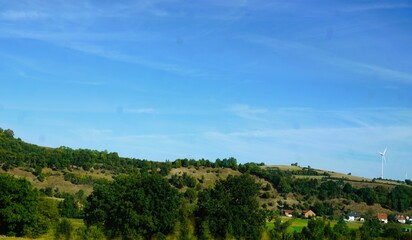Gr&uuml;nes Landschaftspanorama mit Wiesenh&uuml;gel, B&auml;umen, B&uuml;schen Wald und H&auml;usern Tal vor blauem Himmel bei Sonne am Morgen im Sommer