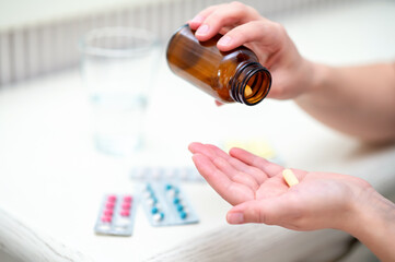 Woman pours pills from amber bottle into her hand with blister packs and a glass of water nearby, concept home medication and daily health routine