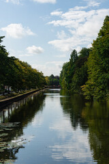 Fototapeta premium Serene canal lined with trees under a bright, partly cloudy sky casting reflections on the water.
