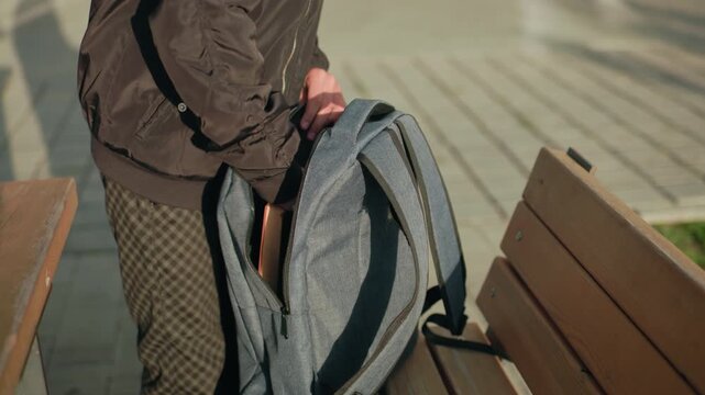Close up of student packing book into backpack while standing beside wooden bench on paved outdoor area with natural sunlight creating strong shadows on ground