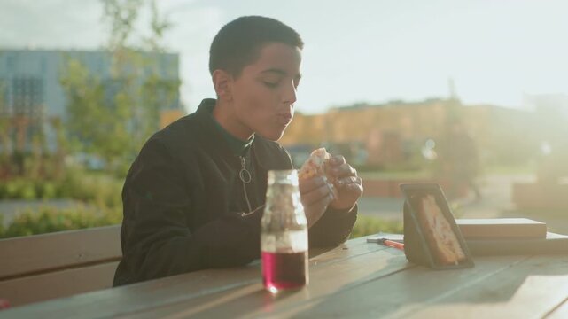 Boy sitting at wooden table outdoors taking bite of sandwich with juice bottle in front and sandwich pack placed nearby under warm daylight in relaxed urban park setting