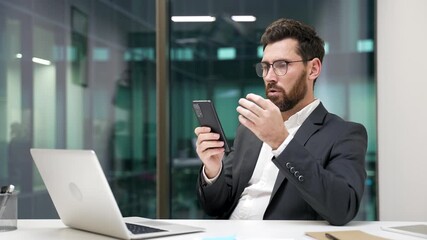 Shocked worried businessman reads bad news on mobile phone while sitting at workplace in a modern business office. Frustrated stressed worker in a formal suit received a negative message on smartphone - Powered by Adobe
