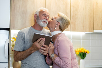 Elderly couple shares a tender moment in a modern kitchen while using a tablet together