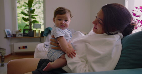 Mother smiling at her baby while holding them on her lap in a sunlit living room, creating a warm...