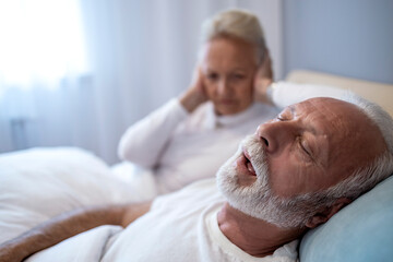 Elderly couple in bed with one partner struggling to sleep due to loud snoring at night in a cozy bedroom setting