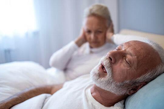Elderly couple in bed with one partner struggling to sleep due to loud snoring at night in a cozy bedroom setting