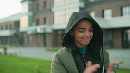 Kid in hooded jacket smiles with joy while holding paper kite outdoors, celebrating playful moment with confidence and excitement against background of urban building and greenery